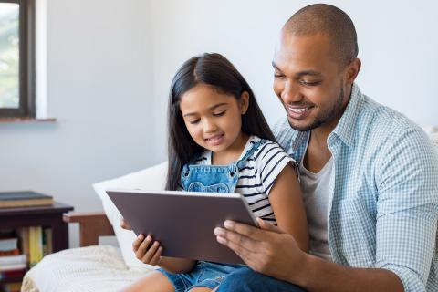 Smiling father holds daughter on lap while they both look at a tablet.
