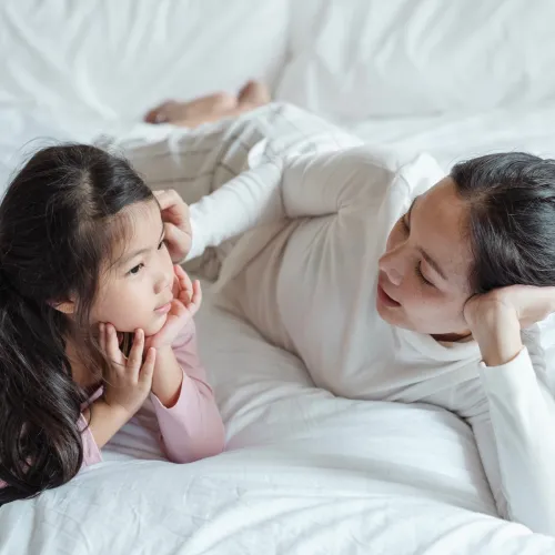 A mother and her daughter have a talk while lying next to each other on a bed with white sheets.