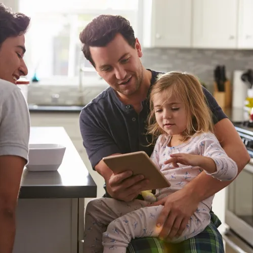 Two dads and their daughter in the kitchen looking at a tablet.