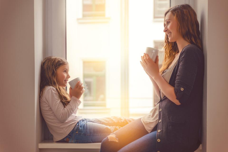 Mother and daughter sit in windowsill while holding mugs